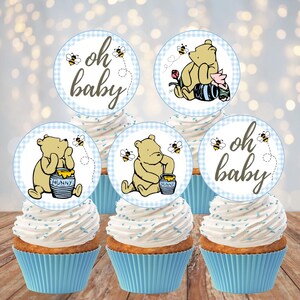 May include: Five cupcakes with light blue liners and white frosting topped with circular paper toppers. The toppers feature illustrations of a yellow bear, honey pots, bees, and the text oh baby in a script font on a light blue checkered background.