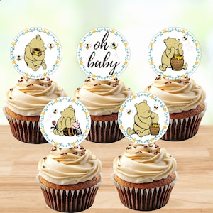 May include: A set of five round cupcake toppers featuring illustrations of a yellow bear with honey pots and bees, framed by a floral wreath. One topper displays the text "oh baby" in black script. Each topper is placed on a frosted cupcake.