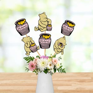 May include: A set of five decorative paper cutouts featuring Winnie the Pooh and honey pots labeled HUNNY, attached to wooden sticks. The cutouts are arranged in a white vase filled with a bouquet of pink, white, and green flowers.