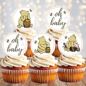 May include: Five cupcakes topped with circular paper decorations featuring illustrations of a yellow bear, a small pink piglet, honey pots labeled 'HUNNY', bees, and the text 'oh baby' in a cursive font, all set against a light beige checkered border.