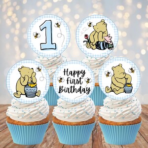 May include: Five cupcakes with light blue liners topped with circular paper toppers featuring Winnie the Pooh, Piglet, bees, the number 1, and the text Happy first Birthday. The toppers have a light blue gingham border and white background.