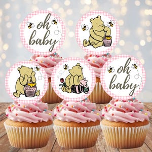 May include: Five cupcakes topped with circular paper toppers featuring Winnie the Pooh characters, honey pots, and bees. The toppers have a pink and white checkered border and include the text "oh baby" in a script font on two of the designs.