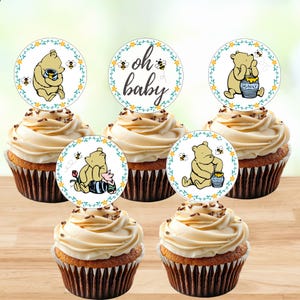 May include: Five cupcakes topped with circular paper decorations featuring Winnie the Pooh illustrations, honey pots, bees, and floral borders. One topper displays the text oh baby in a cursive script, while others show Pooh bear in various poses.