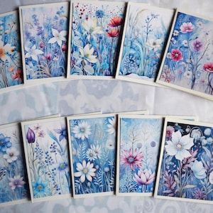 May include: A set of ten floral greeting cards. Each card displays a watercolour painting of diverse flowers in blue, white, pink, and purple hues. The cards have a white border and are arranged on a white and blue patterned surface.