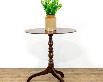 May include: A round dark brown wooden pedestal table with a central turned column and three curved legs. A cylindrical tan ceramic jar sits on the tabletop, containing a cluster of bright green artificial leafy stems and foliage.