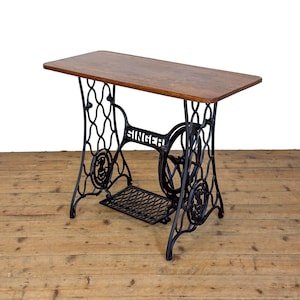 May include: A rectangular wooden table top supported by a black cast iron Singer sewing machine base. The base features intricate scrollwork, a central treadle pedal, and a large side wheel with the brand name Singer clearly visible in white.