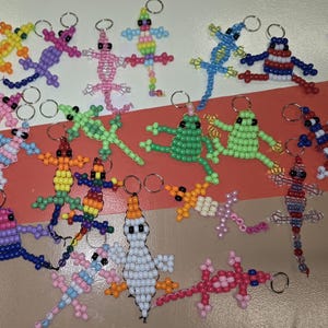 May include: A collection of handmade beaded keychains shaped like lizards and frogs in various colors including green, blue, pink, yellow, and purple. Each small animal figure is constructed from plastic pony beads and attached to a metal ring.