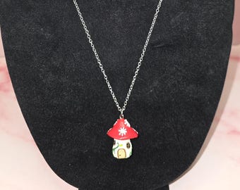 May include: A silver-toned metal chain necklace featuring a pendant shaped like a mushroom house. The mushroom cap is red with white floral accents, while the stem is white with a small arched door and green vine details painted on the surface.