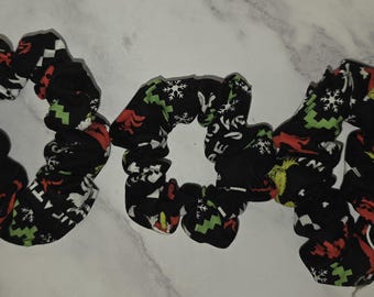 May include: Three black fabric hair scrunchies featuring a repeating holiday-themed print. The pattern includes green cartoon characters, red shapes, white snowflakes, and geometric designs. Each scrunchie has a gathered, elasticized construction.