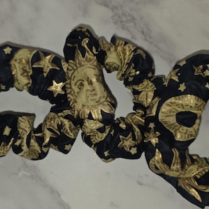 May include: Two fabric hair scrunchies featuring a black background with a repeating gold metallic print of sun faces, crescent moons, and various stars. The scrunchies have a gathered, elasticised design with a soft, pleated texture throughout.