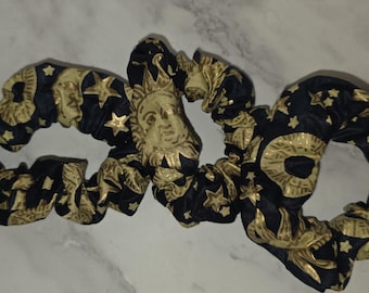 May include: Two fabric hair scrunchies featuring a black background with a repeating gold metallic print of sun faces, crescent moons, and various stars. The scrunchies have a gathered, elasticized design with a soft, pleated texture throughout.