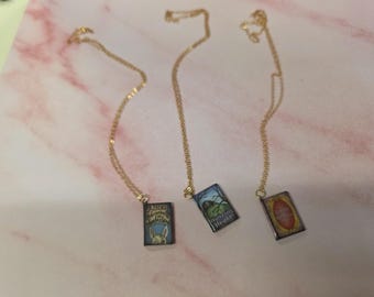 May include: Three gold-tone chain necklaces, each featuring a rectangular pendant designed to look like a miniature book cover. The pendants display the titles Alice's Adventures in Wonderland, Wuthering Heights, and The Little Witch, with colorful illustrations on each.