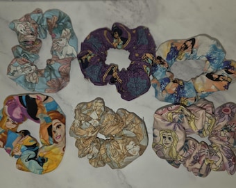 May include: A set of six fabric hair scrunchies featuring various cartoon character prints. The collection includes patterns with light blue, purple, multi-colored, yellow, tan, and pink backgrounds, each displaying different animated figures.