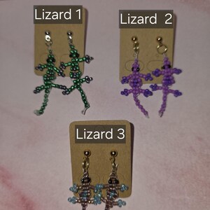 May include: Three pairs of lizard-shaped beaded earrings displayed on brown cardboard cards labelled Lizard 1, Lizard 2, and Lizard 3. The first pair is green, the second is purple, and the third is a mix of brown and blue beads with gold-tone posts.