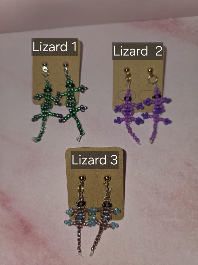 May include: Three pairs of lizard-shaped beaded earrings displayed on brown cardboard cards labelled Lizard 1, Lizard 2, and Lizard 3. The first pair is green, the second is purple, and the third is a mix of brown and blue beads with gold-tone posts.