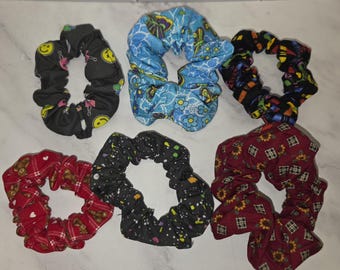 May include: A collection of six fabric hair scrunchies featuring various patterns: a black one with yellow smiley faces, a blue one with butterflies and flowers, a black one with colorful shapes, a red one with teddy bears, a black one with paint splatters, and a maroon one with sunflowers.