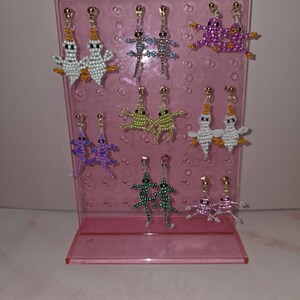 May include: A pink acrylic display stand holding seven pairs of beaded earrings. The earrings feature various animal shapes including ghosts, lizards, and frogs in colours such as white, purple, green, and pink, all attached to gold-toned metal posts.