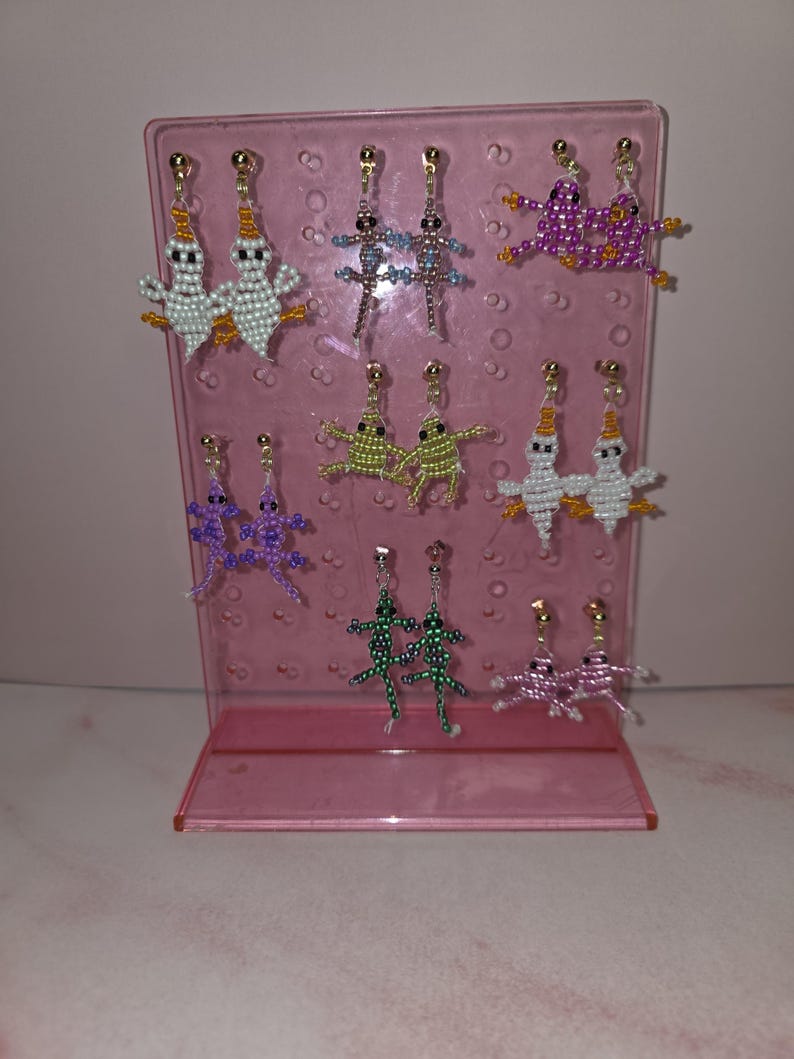 May include: A pink acrylic display stand holding seven pairs of beaded earrings. The earrings feature various animal shapes including ghosts, lizards, and frogs in colours such as white, purple, green, and pink, all attached to gold-toned metal posts.