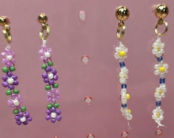 May include: Two pairs of beaded dangle earrings featuring gold-tone ball studs. One pair displays purple and green flower patterns with white centers, while the other pair features white flower patterns with yellow centers and blue spacer beads.