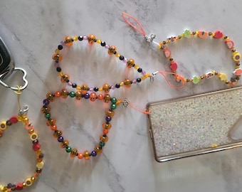 May include: Three beaded phone charms and lanyards featuring colorful plastic beads, including orange pumpkin-shaped beads, small round metallic beads, and fruit-shaped charms. The charms are attached to a smartphone case and a car key fob.
