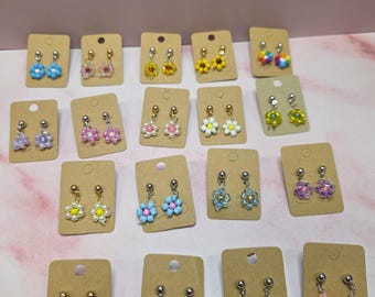 May include: A collection of nineteen pairs of flower-shaped beaded earrings, each mounted on a small rectangular brown card. The earrings feature various colors including blue, pink, yellow, white, purple, green, and multicolored beads with metal posts.