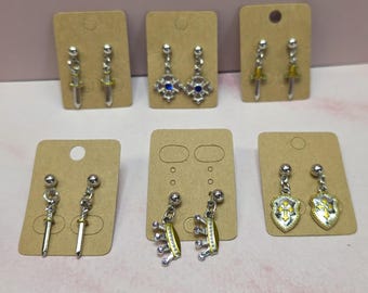 May include: Six pairs of silver-toned metal drop earrings displayed on individual brown cardboard cards. The designs include swords, a blue-centered star, crowns, and shields with gold-colored accents, all featuring a post-back closure style.