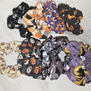 May include: A collection of eight fabric hair scrunchies featuring various patterns including white spiderwebs on black, orange and black plaid, candy corn, orange pumpkins on black, autumn leaves, metallic silver, and purple with black cats.