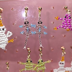 May include: A pair of beaded lizard earrings featuring metallic brown bodies with light blue accents and black bead eyes. The lizards are suspended from gold-toned metal earring posts. The design is crafted with small seed beads in a detailed pattern.