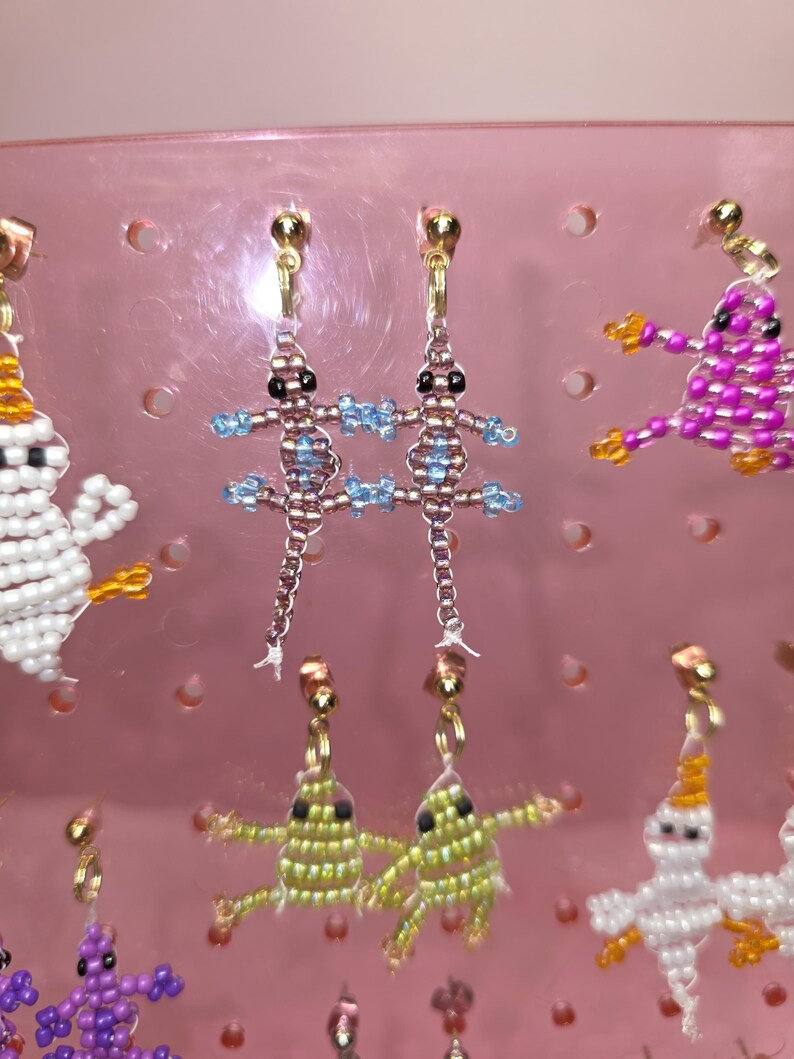 May include: A pair of beaded lizard earrings featuring metallic brown bodies with light blue accents and black bead eyes. The lizards are suspended from gold-toned metal earring posts. The design is crafted with small seed beads in a detailed pattern.