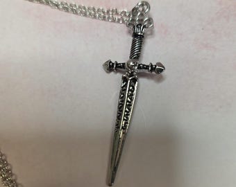 May include: A silver-toned metal pendant shaped like a dagger, featuring intricate engraved patterns along the blade and a twisted handle design. The pendant is attached to a delicate silver-toned chain link necklace that extends from the top.