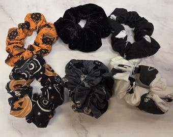 May include: A collection of six fabric hair scrunchies featuring various designs. One is orange with black cats, one is solid black velvet, one is black with white ghosts, one is black with pumpkins and stars, one is grey with cats, and one is white with black shapes.