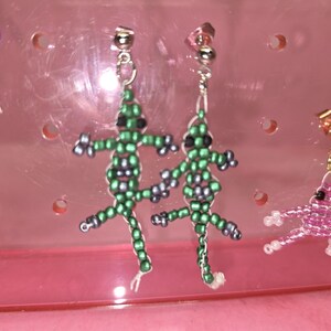 May include: A pair of dangle earrings featuring lizard-shaped charms crafted from small green and grey seed beads. The lizards have black bead eyes and are attached to silver-toned metal stud posts. The earrings are displayed on a pink plastic stand.
