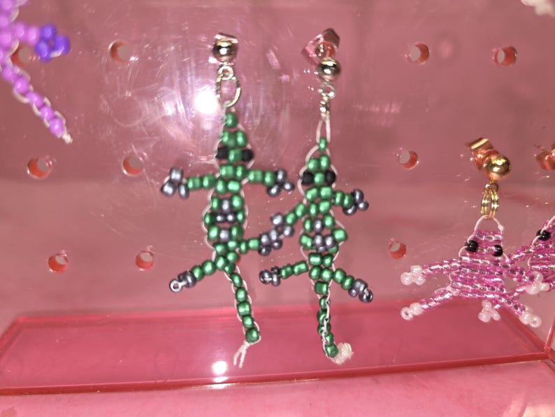 May include: A pair of dangle earrings featuring lizard-shaped charms crafted from small green and grey seed beads. The lizards have black bead eyes and are attached to silver-toned metal stud posts. The earrings are displayed on a pink plastic stand.