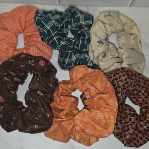 May include: A collection of six fabric hair scrunchies featuring various patterns and colours, including an orange polka dot, a dark green plaid, a cream mushroom print, a brown floral print, an orange wavy line print, and a black geometric pattern.