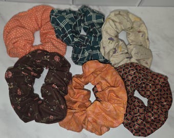 May include: A collection of six fabric hair scrunchies featuring various patterns and colors, including an orange polka dot, a dark green plaid, a cream mushroom print, a brown floral print, an orange wavy line print, and a black geometric pattern.