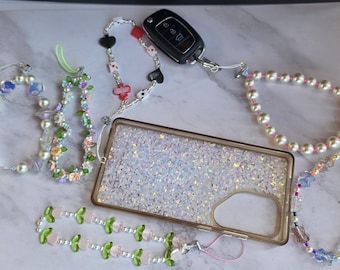 May include: A rectangular phone case with a shimmering iridescent glitter back and gold frame, surrounded by six decorative beaded phone charms featuring various designs including pearls, floral motifs, playing card suits, and green leaf shapes.