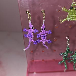 May include: A pair of dangle earrings shaped like lizards, crafted from small purple and light purple beads with black bead eyes. The earrings feature gold-toned metal posts and are attached to a translucent pink plastic display stand with holes.