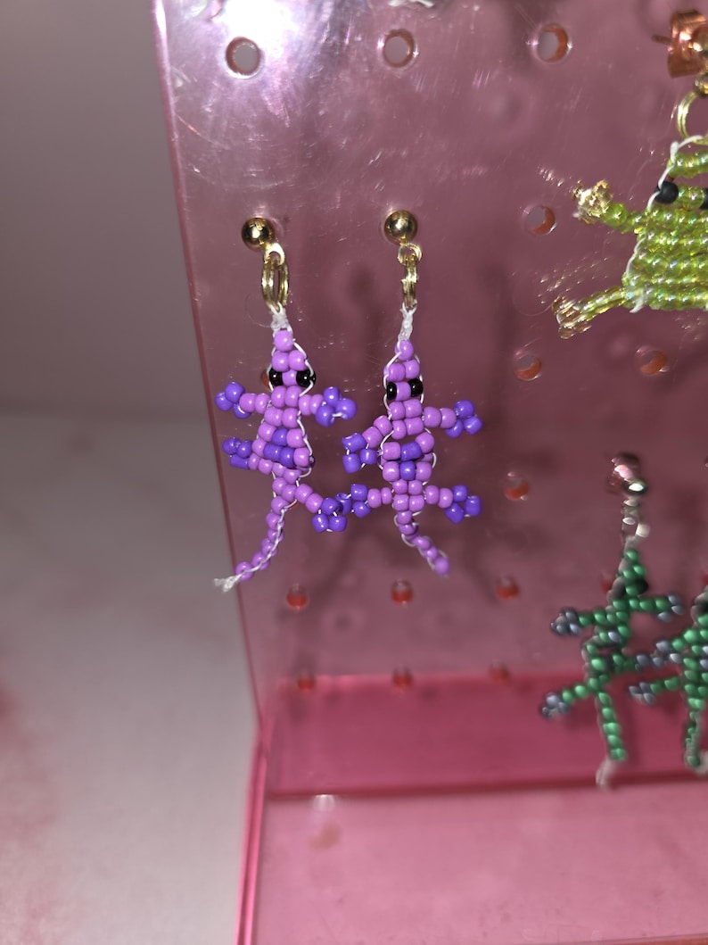 May include: A pair of dangle earrings shaped like lizards, crafted from small purple and light purple beads with black bead eyes. The earrings feature gold-toned metal posts and are attached to a translucent pink plastic display stand with holes.