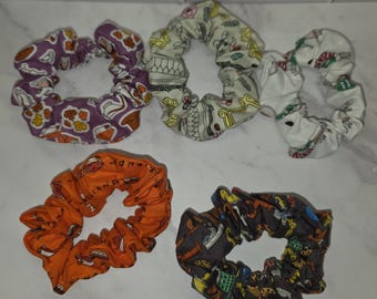 May include: Five fabric hair scrunchies featuring various patterns. One is purple with autumn leaves and food, one is cream with yellow and black line art, one is white with green and red motifs, one is orange with text, and one is dark gray with icons.