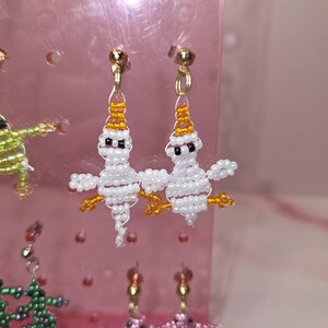 May include: A pair of handmade beaded earrings featuring white ghost-shaped figures with orange bead accents on the head and feet, black bead eyes, and gold-toned metal earring hooks, displayed on a pink plastic jewellery stand.