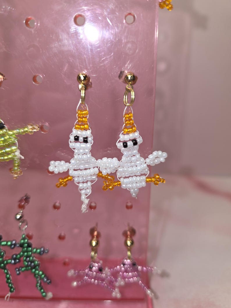 May include: A pair of handmade beaded earrings featuring white ghost-shaped figures with orange bead accents on the head and feet, black bead eyes, and gold-toned metal earring hooks, displayed on a pink plastic jewellery stand.