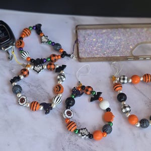 May include: Three beaded wristlet lanyards featuring Halloween-themed charms including ghosts, bats, pumpkins, and eyeballs, interspersed with orange, black, and white patterned beads. One lanyard is attached to a car key fob and a glittery phone case.