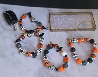 May include: Three beaded wristlet lanyards featuring Halloween-themed charms including ghosts, bats, pumpkins, and eyeballs, interspersed with orange, black, and white patterned beads. One lanyard is attached to a car key fob and a glittery phone case.