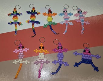 May include: A collection of nine lizard-shaped keychains crafted from colorful plastic pony beads. Each lizard features a distinct color pattern, including rainbow, pastel pink, blue, orange, white, and purple, all attached to a silver metal ring.