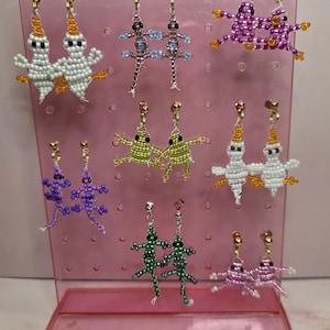Puede incluir: Una colección de ocho pares de pendientes colgantes de cuentas exhibidos en un soporte de plástico rosa. Los pendientes presentan varias formas de animales, incluyendo fantasmas blancos, lagartos morados y verdes, y ranas rosas y verdes, hechos con pequeñas cuentas.