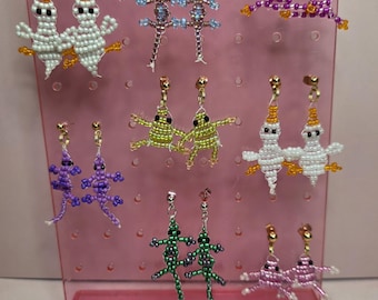 May include: A collection of eight pairs of beaded dangle earrings displayed on a pink plastic stand. The earrings feature various animal shapes including white ghosts, purple and green lizards, and pink and green frogs, all crafted with small colorful beads.