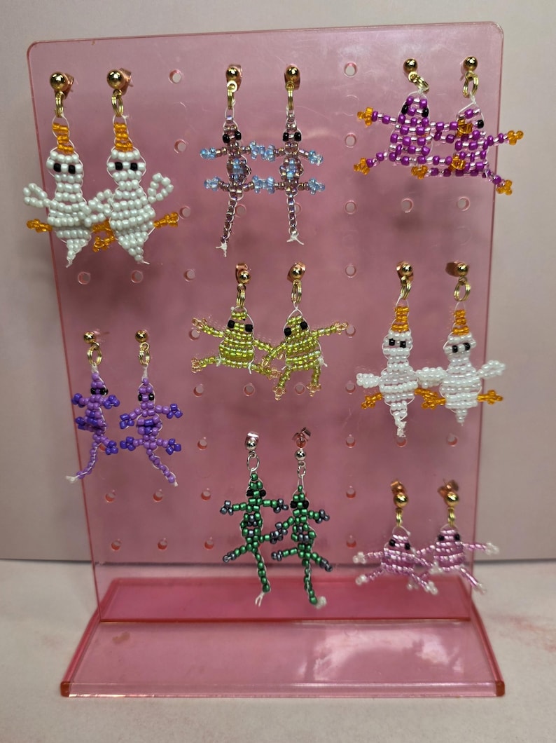 May include: A collection of eight pairs of beaded dangle earrings displayed on a pink plastic stand. The earrings feature various animal shapes including white ghosts, purple and green lizards, and pink and green frogs, all crafted with small colourful beads.