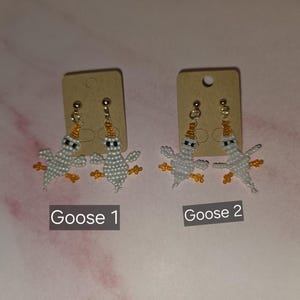 May include: Two pairs of beaded earrings shaped like geese, each featuring white bodies, orange beaks, and orange feet. The earrings are attached to brown cardboard display cards labelled Goose 1 and Goose 2, with gold-toned metal earring posts.