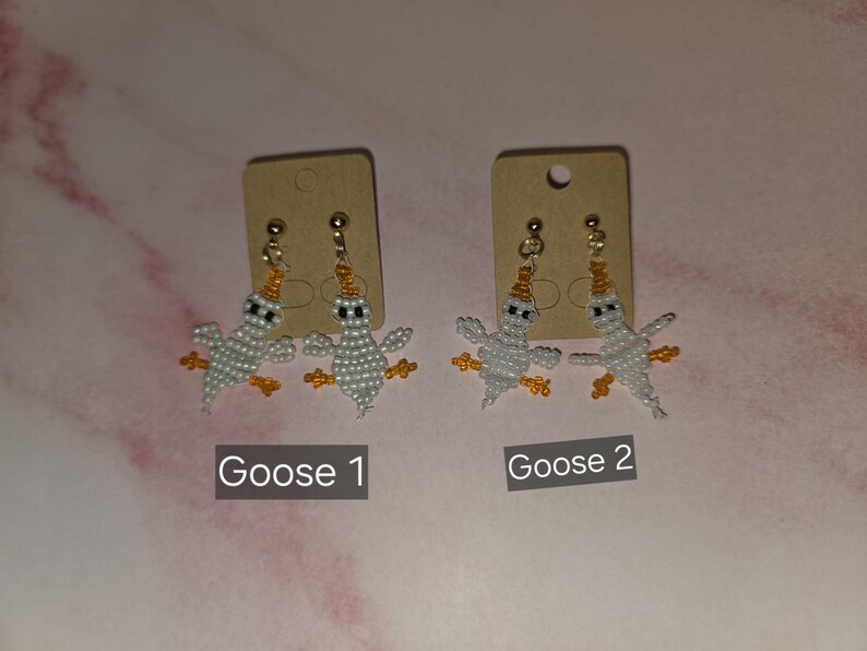 May include: Two pairs of beaded earrings shaped like geese, each featuring white bodies, orange beaks, and orange feet. The earrings are attached to brown cardboard display cards labelled Goose 1 and Goose 2, with gold-toned metal earring posts.