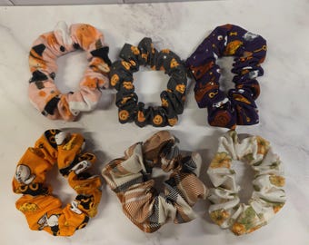 May include: A set of six fabric hair scrunchies featuring various patterns including orange pumpkins on a light pink background, jack-o'-lanterns on dark gray, purple with Halloween motifs, orange with ghosts, a brown and cream plaid, and white with pumpkins.